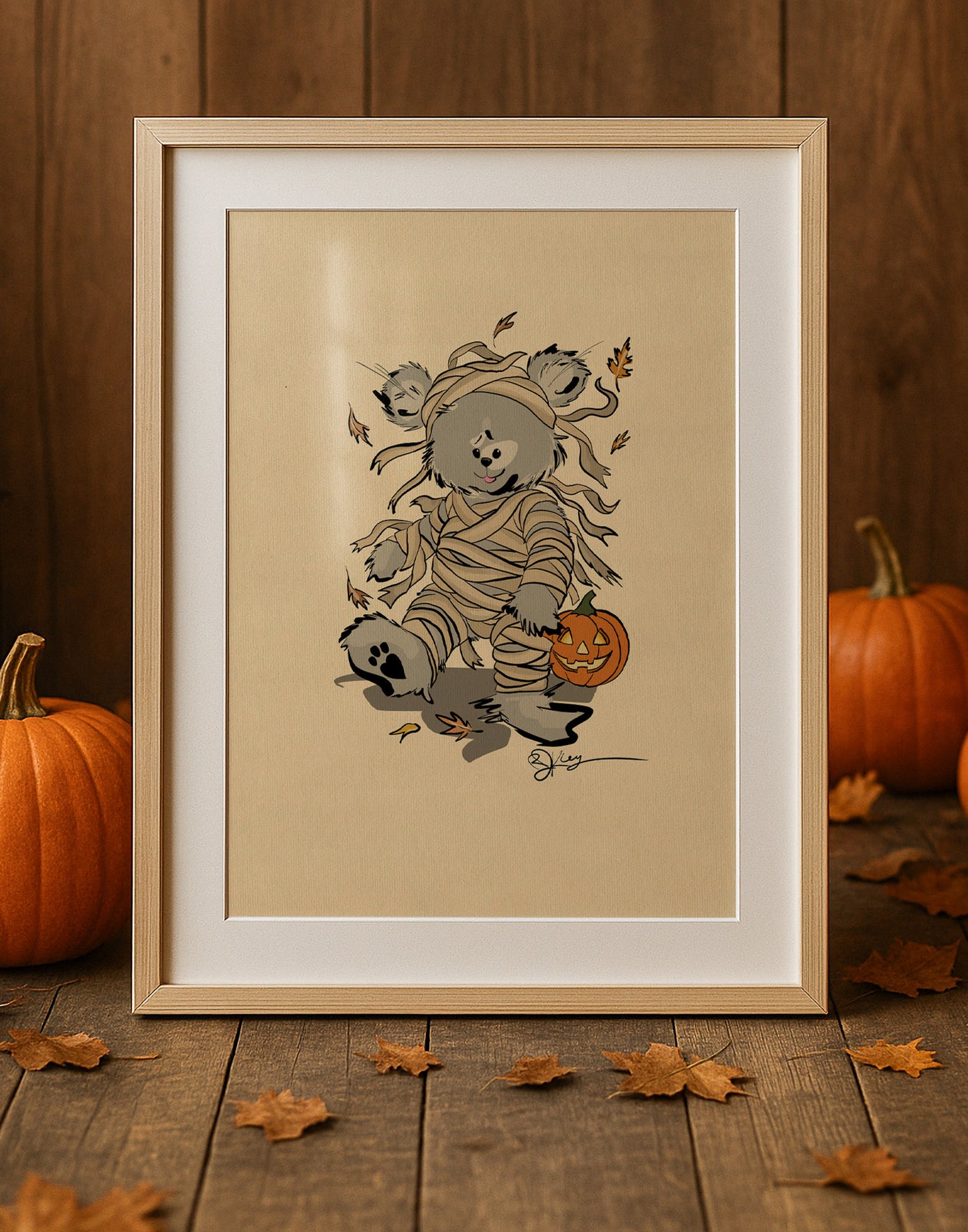 A framed print from The Pola Bears Halloween 2025 Collection, featuring the limited edition print Boris. The print shows a Pola Bear wrapped in bandages like a mummy, with a carved pumpkin. The framed print is placed on a rustic wooden table with autumn leaves scattered around it and two pumpkins on either side.