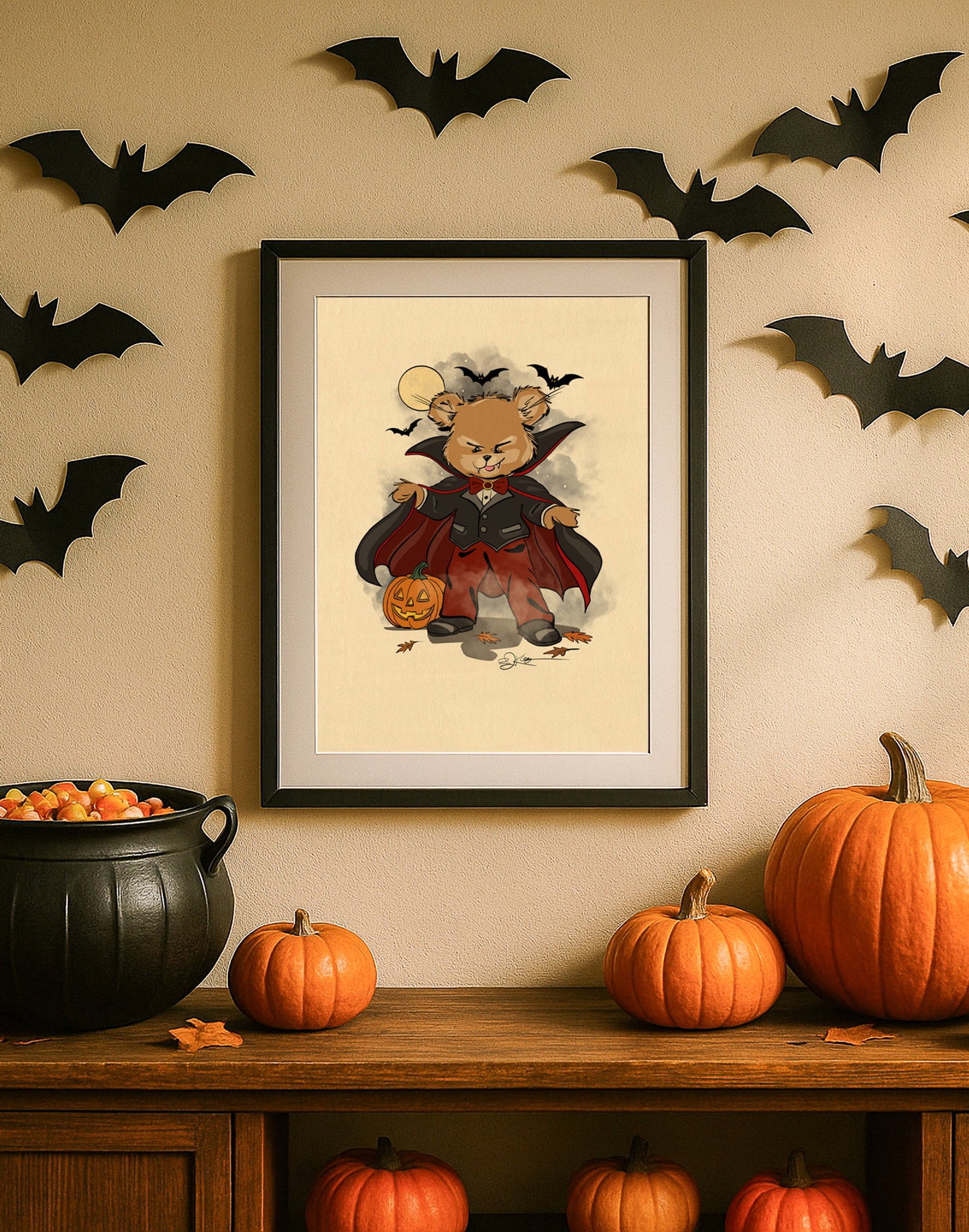 A framed print from The Pola Bears Halloween 2025 Collection, featuring the limited edition print Christopher. The print shows a Pola Bear dressed as a vampire, holding a carved pumpkin with bats flying around its head. The framed print sits on a wooden cabinet next to a black cauldron filled with candy corn, pumpkins, and a string of paper bats decorating the wall behind it.