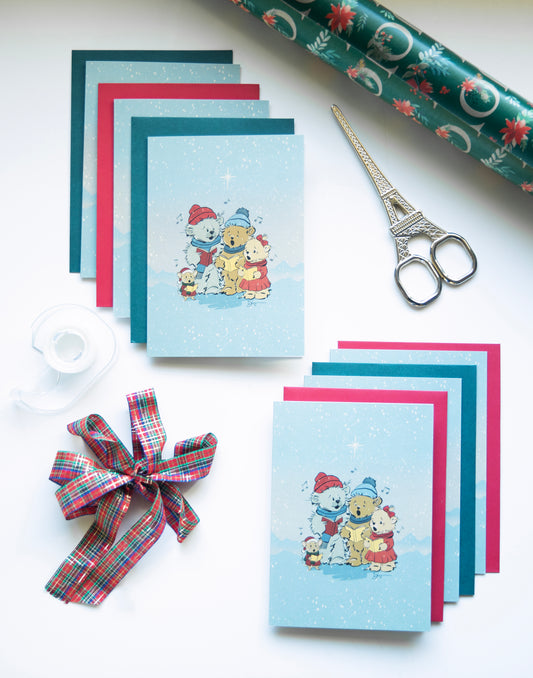 Flat lay photography of "The Carolers" vintage-style Christmas greeting cards by The Pola Bears. Two stacks of light blue cards featuring an illustration of singing Pola Bears are fanned out with matching red and green envelopes. The festive scene includes a roll of green poinsettia wrapping paper, silver scissors, clear tape, and a plaid ribbon bow on a white background.