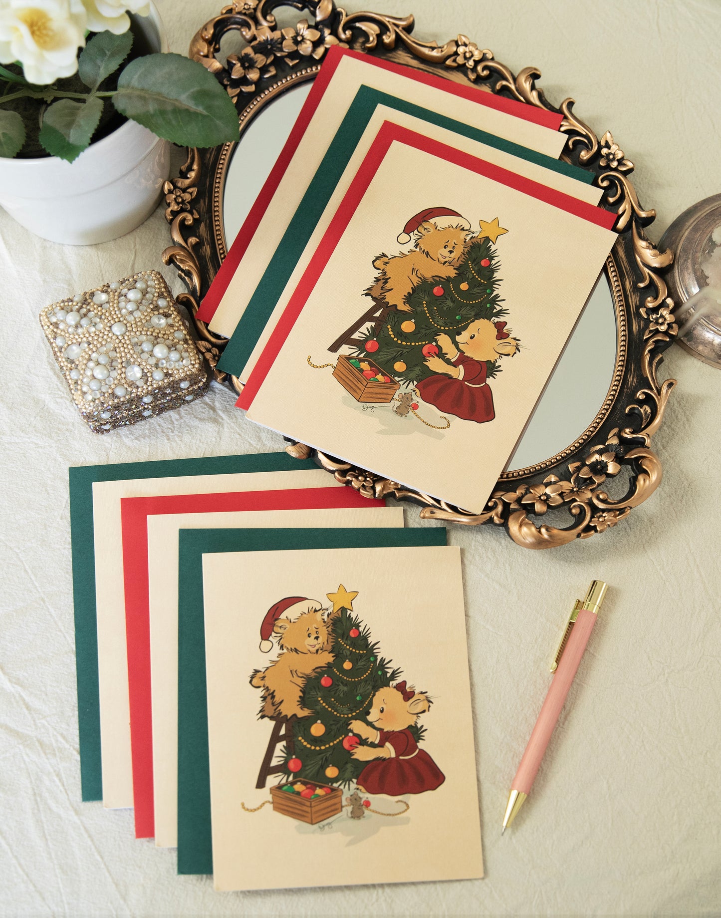 Flat lay of vintage-inspired Christmas greeting cards by The Pola Bears. The hand-drawn illustration features two bears and a small mouse decorating a Christmas tree. The cards are displayed with matching red and green envelopes on an ornate gold mirror tray with a pearl box and pink pen.