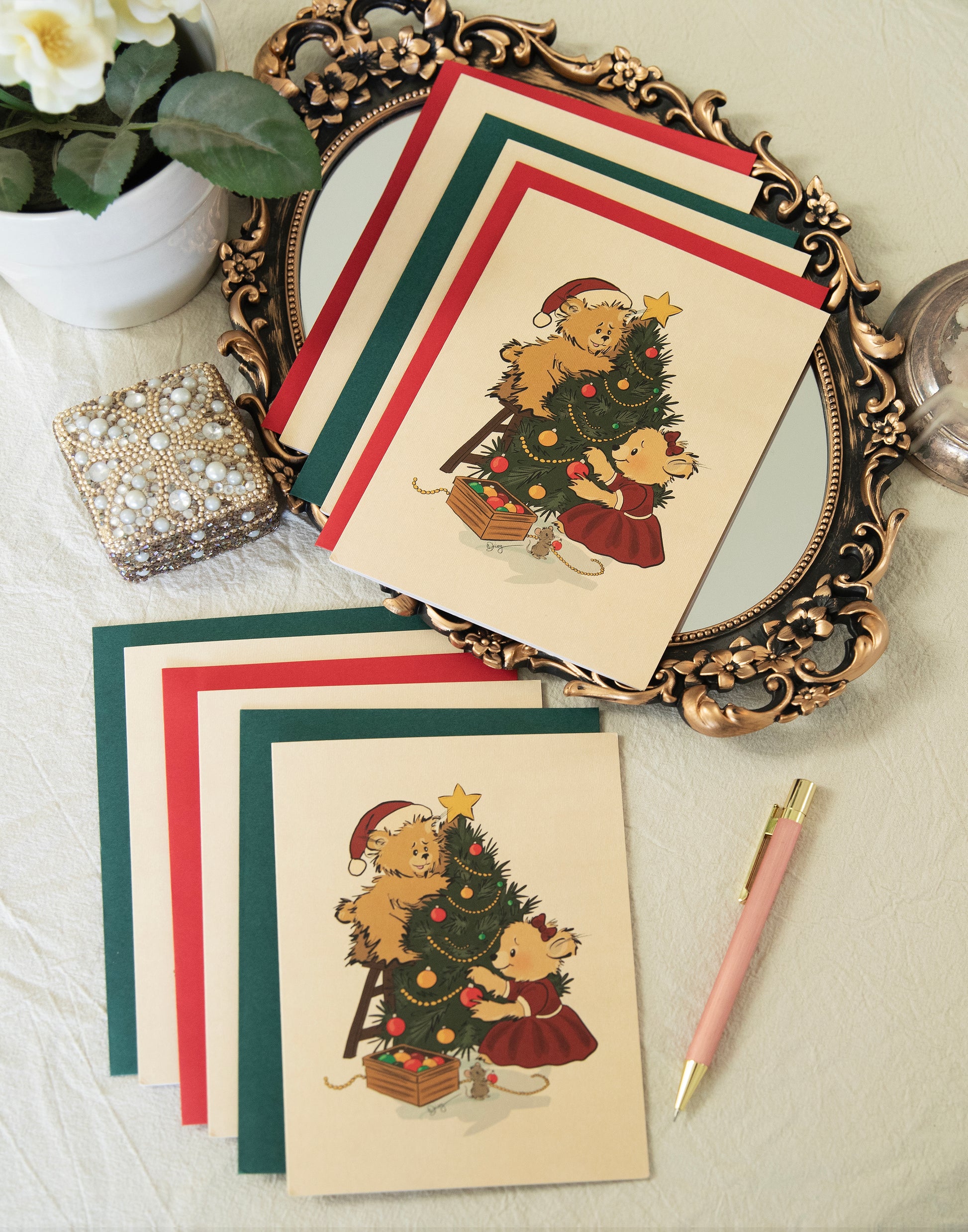 Flat lay of vintage-inspired Christmas greeting cards by The Pola Bears. The hand-drawn illustration features two bears and a small mouse decorating a Christmas tree. The cards are displayed with matching red and green envelopes on an ornate gold mirror tray with a pearl box and pink pen.