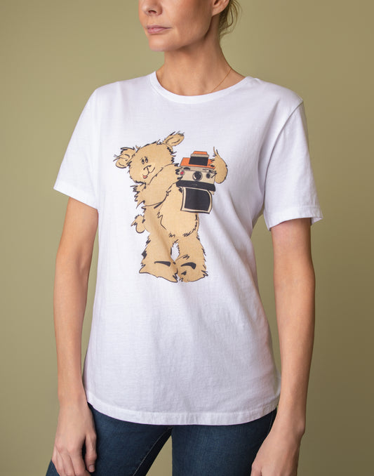 A woman wearing a white short-sleeve t-shirt featuring a whimsical illustration of Greg the Pola Bear holding a vintage camera.