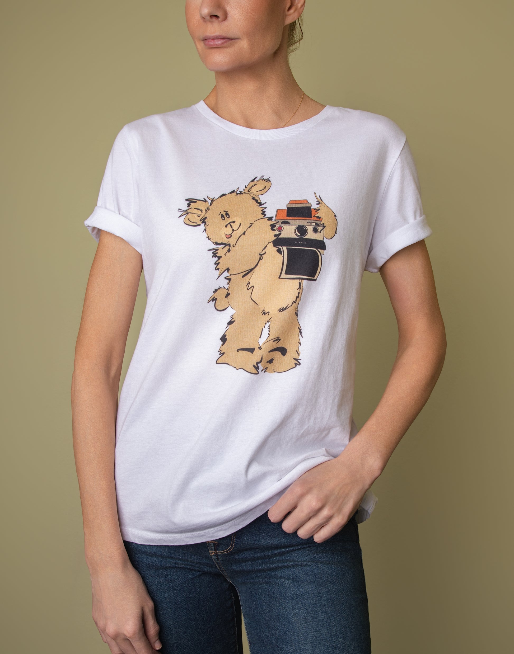 Model wearing a white graphic tee styled with rolled sleeves, featuring the 'Greg' vintage Pola Bear illustration. She poses casually with one hand in her blue jeans pocket against an olive green background.